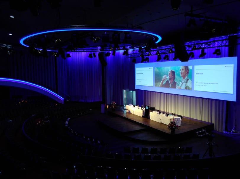 Grosser Höhrsaal mit beleuchteter Bühne auf der ein Podium steht und darüber eine grosse Leinwand mit drei Folien angezeigt wird