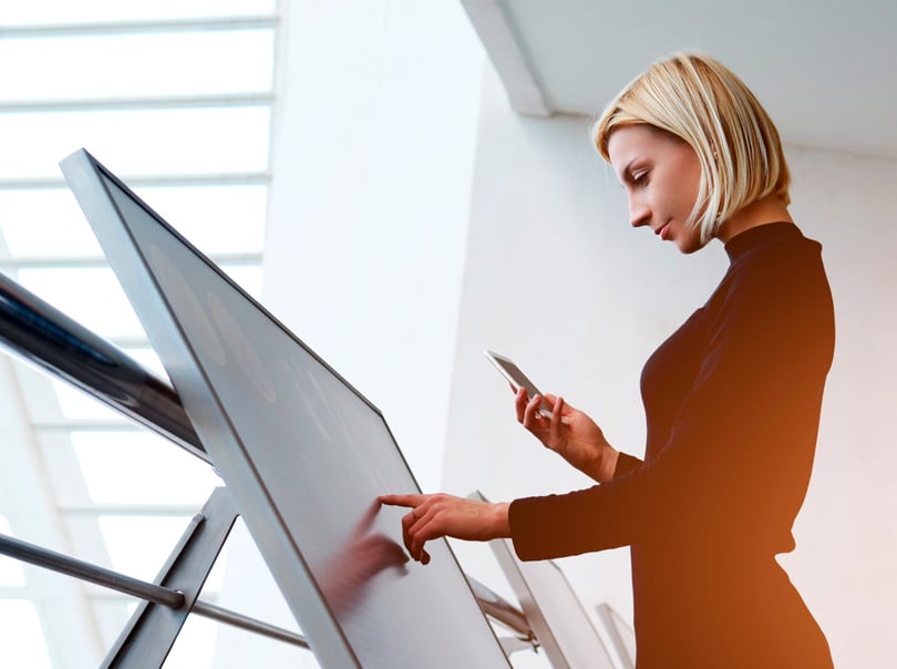Eine Mitarbeiterin drückt auf einen Monitor mit Touchfunktion, der als Digital Signage-Lösung Informationen abbildet.
